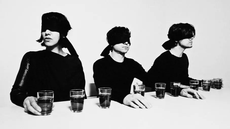 Three blindfolded individuals seated at a table, each with multiple glasses of liquid arranged in front of them.