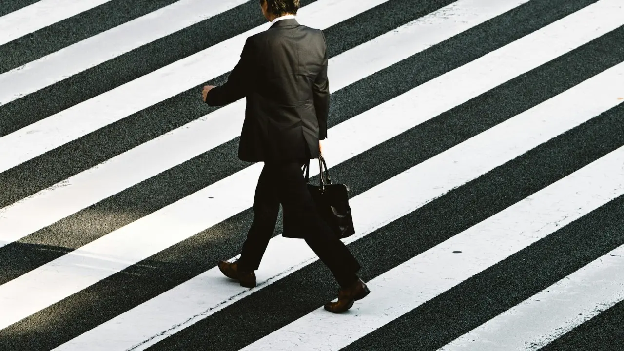 A man in a business suit walks across a wide black-and-white striped crosswalk, carrying a briefcase.