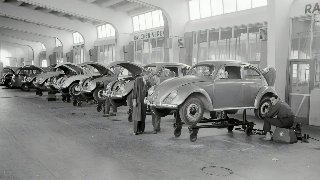 Historic black-and-white photo of a Volkswagen factory line, with multiple VW Beetles being assembled and workers inspecting and repairing the cars.