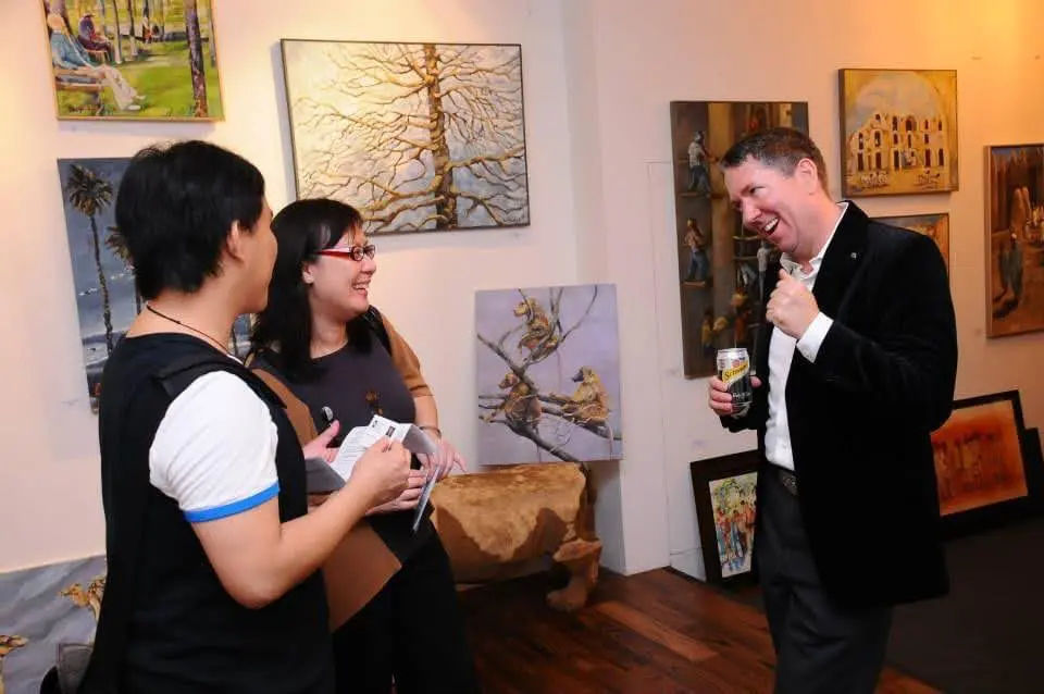 Guests and gallery owner talking at an art exhibition in Singapore, surrounded by paintings displayed on the walls.